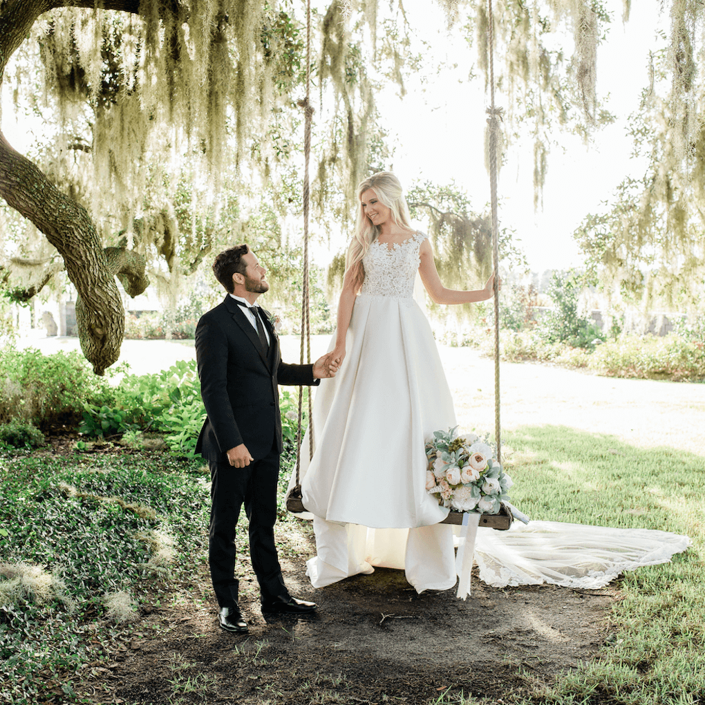 Groom stands on the ground next to his bride who is standing on a swing in her wedding dress. They're holding hands and Spanish moss hangs from a tall tree behind them. The whole photo is full of beautiful natural light.