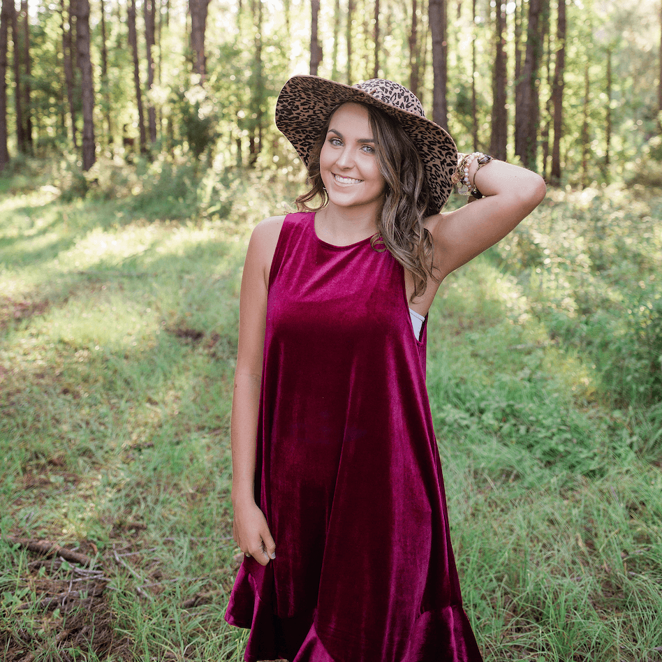 A high school senior poses for senior portraits wearing a leopard print hat and a red velvet dress. The photo is outside with bright natural light pouring through the trees behind the subject.