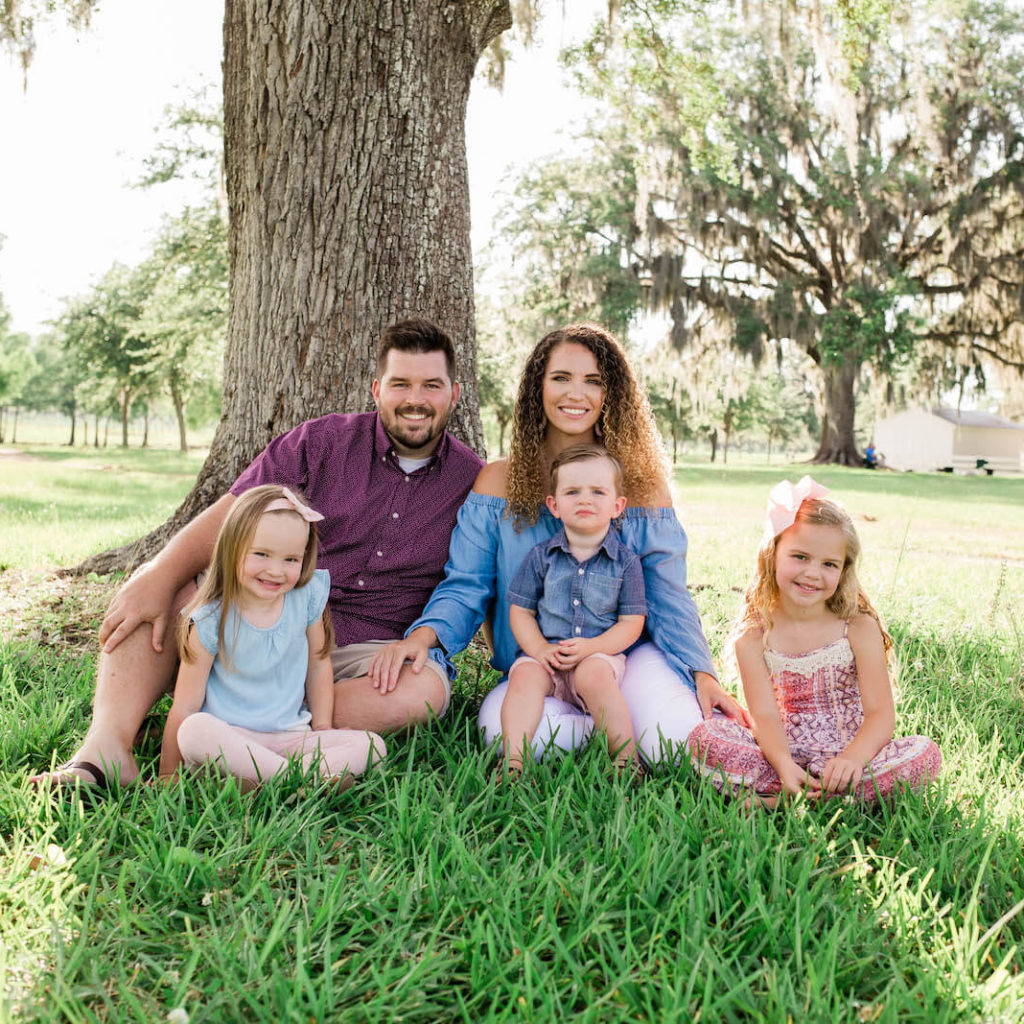 A family (mom, dad, two daughters and son) pose for a family portrait in a grassy field in front of a large tree.