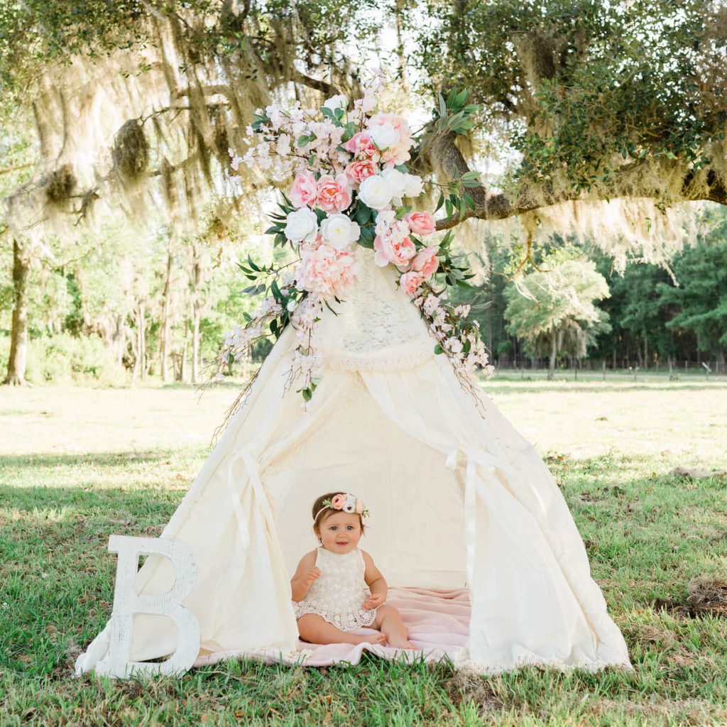 A baby girl wearing a floral headband sits in a white formal tent with flowers on top of it outside under a tree full of Spanish moss.